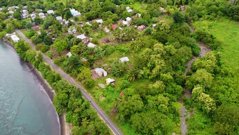 Aerial-drone-of-motorbike-riding-along-coastal-road-alongside-beautiful-ocean,-local-community-houses-and-green-tree-landscape-in-Alor-Island,-East-Nusa-Tenggara,-Indonesia