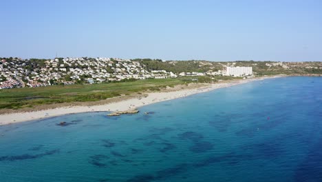 Menschen-Entspannen-Sich-Am-Strand-Von-Son-Bou-Auf-Menorca,-Spanien,-Mit-Kristallklarem,-Blauem-Wasser-Und-Luxusimmobilien-Im-Hintergrund