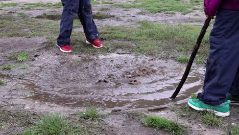 Two-ethnically-diverse-children-playing-with-sticks-in-a-muddy-puddle