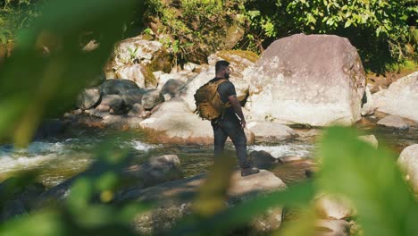 Slow-motion-young-male-hiker-along-mountain-river-surounded-by-boulders-and-trees,-looking-around-during-hike-in-jungle