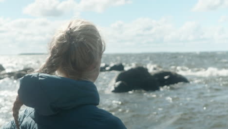Close-up-of-Blonde-Girl-Standing-on-the-beach-by-the-Sea,-Looking-out-to-the-waves,-Two-Kayaks-Laying-on-the-Beach,-Windy-Cold-Summer-Day-in-Finland,-Near-Vaasa