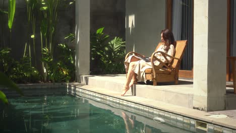 Expat-woman-working-online-in-her-luxury-home-with-swimming-pool-during-sunny-day-and-using-laptop-computer-to-work-from-home-early-morning,-female-professional-lady-using-technology-doing-remote-job