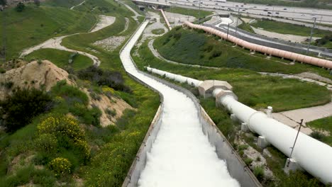 aerial-drone-los-angeles-aqueduct-cascades-performing-flood-control-by-releasing-water-down-green-hill-with-flowers-and-cars-driving-by