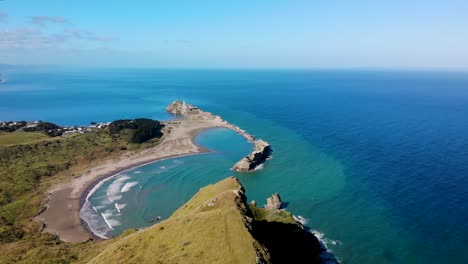 Fly-over-rocky-cliff-to-the-amazing-scenic-spot,-sandy-beach,-bay-surrounded-by-reef-and-iconic-New-Zealand-lighthouse
