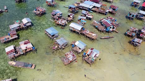 Revealing-aerial-shot-of-a-boat-driving-in-a-water-village-of-the-Bajau-Laut-with-water-houses-and-crystal-clear-sea,-at-Omadal-Semporna,-Sabah,-Malaysia