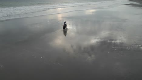 Graue-Wolken-Spiegeln-Sich-Am-Nassen-Sandstrand,-Während-Ein-Mann-Auf-Einem-Motorrad-Aufsteht