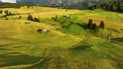 Aerial-view-over-sunlit-meadows,-revealing-a-rainbow-in-front-of-Alpe-di-Siusi-peaks,-in-Dolomites,-Italy