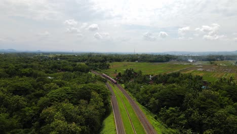 Drone-view-following-train-crossing-bridge-in-tropical-countryside,-Indonesia