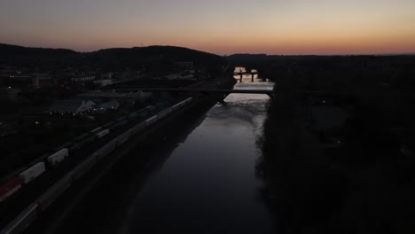 An-aerial-view-of-the-Lehigh-River-in-Bethlehem,-Pennsylvania,-after-sunset-with-long-cargo-trains-on-the-left-of-the-shot