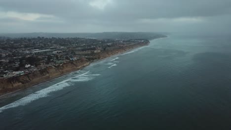 View-from-a-drone-tilting-down-over-the-ocean-showing-the-sea,-the-beach-and-a-town-close-to-the-edge-of-a-cliff