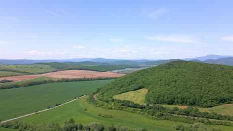 Grüne-Felder-Und-Ackerland-In-Der-Mittagssonne,-Aufgenommen-Mit-Einer-Drohne-Mit-Einer-Hügeligen-Landschaft-Im-Hintergrund