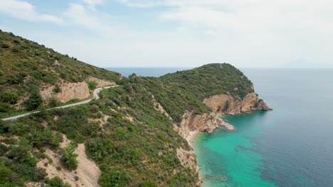 Aerial-View-Of-A-Rocky-Seaside,-Road-With-A-Car-Passing-By,-Turquoise-Water,-Lush-Vegetation,-Fari-Beach,-Thassos-Island,-Greece