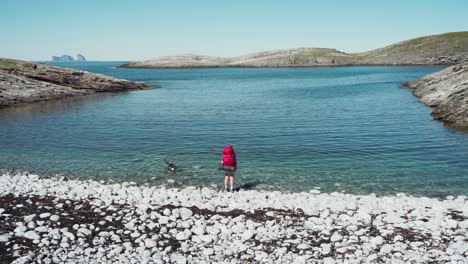 Rückansicht-Eines-Männlichen-Rucksacktouristen,-Der-Seinen-Hund-Beim-Schwimmen-Am-Strand-Der-Insel-Lovund-In-Norwegen-Beobachtet