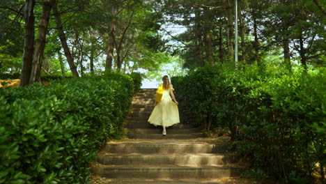 Rear-View-of-Blonde-Woman-in-Yellow-Summer-Dress-with-Orange-Eco-Bag-Walking-Upstairs-in-Green-Lush-Park-in-Slow-Motion