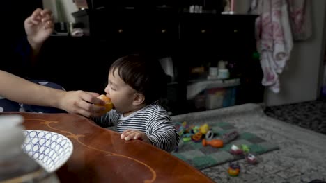 Adorable-8-Month-Old-Indian-Baby-Being-Fed-Orange-Held-By-Mother-As-He-Stands-Beside-Table-Indoors