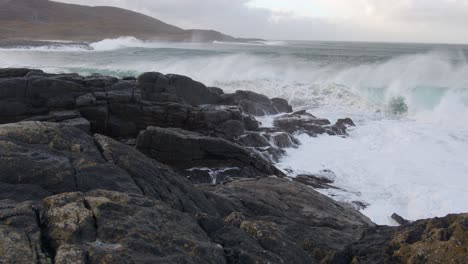 Slow-motion-shot-of-large,-white-waves-breaking-over-a-cliffside-during-a-storm-in-the-bay-by-Tangasdale-beach,-near-Castlebay-on-the-Isle-of-Barra