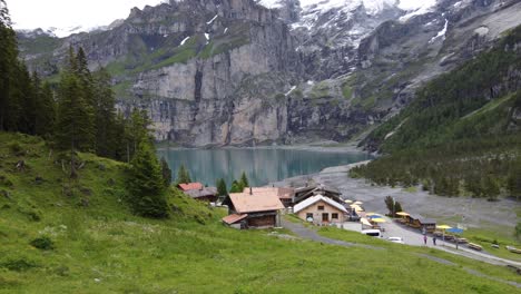 Toma-Aérea-Hacia-Atrás:-Vista-De-2-Excursionistas-En-Su-Camino-A-La-Cafetería-De-La-Montaña-Y-Montañas-Gigantes-Bluemlisalp-Que-Rodean-El-Lago-Glaciar-Turquesa-Szure-Oeschinensee-En-Kandersteg,-Suiza