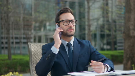 Businessman-talks-on-phone-writes-notes-sitting-at-the-park-in-front-of-office