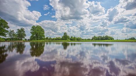 Timelapse-shot-of-white-cloud-movement-over-pristine-lake-surrounded-by-green-vegetation-along-rural-countryside-throughout-the-day
