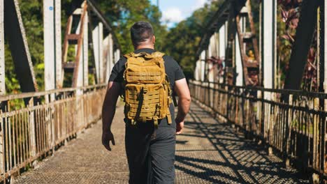 Young-man-traveler-with-backpack-walking-on-rusty-old-bridge-outdoors-on-sunny-summer-day