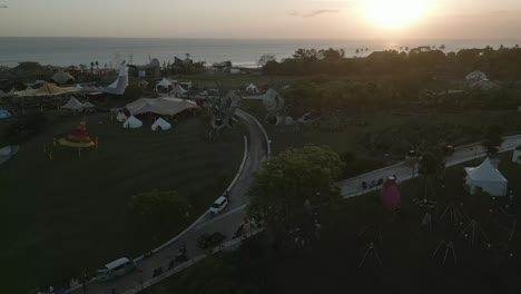 Sunset-flyover:-tents-and-sculptures-in-coastal-Bali-park,-Indonesia