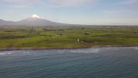 Aerial-scenic-of-New-Zealand-coastal-landmark-with-lighthouse,-iconic-Taranaki-volcano-on-backdrop