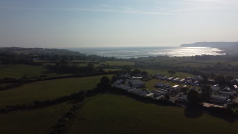 Aerial-view-Welsh-caravan-park-at-sunrise-overlooking-shimmering-coastal-bay-and-rolling-countryside-hills,-panning-left