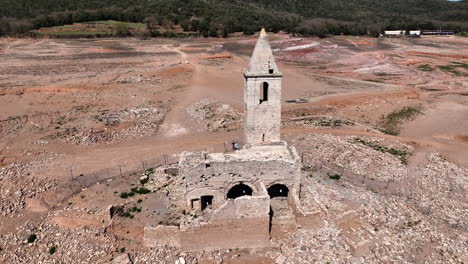 Aerial-view-orbiting-sunlit-abandoned-Church-of-Sant-Roma,-Catalan-tower-on-deserted-rocky-coastline-of-the-Sau-reservoir