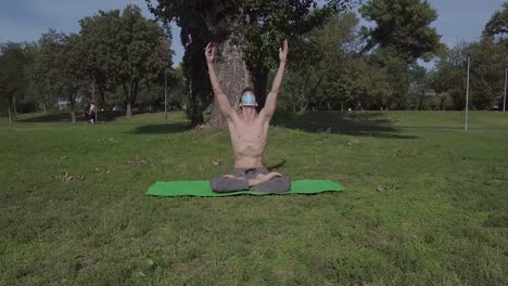 Sporty-young-man-with-the-mask-doing-yoga-in-the-park-on-the-green-mat,-yogi-performs-yoga-stretching-in-nature