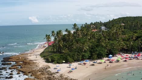 Left-trucking-aerial-drone-shot-of-the-popular-tropical-Coquerinhos-beach-surrounded-by-palm-trees-and-covered-in-umbrellas-and-small-waves-crashing-into-exposed-rocks-in-Conde,-Paraiba,-Brazil