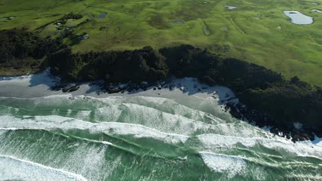 Colourful-aerial-scene-of-ocean-waves-and-white-sand-beach,-green-pastures-coastal