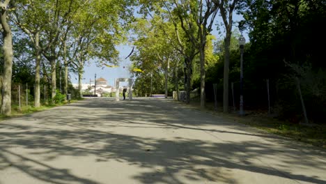 Joven-Con-Mochila-De-Pie-Y-Posando-En-Medio-De-La-Carretera-Del-Parque-En-El-Palacio-Nacional-De-Montjuic,-Palau-Nacional-En-Barcelona,-España-En-Un-Hermoso-Día-Soleado-Con-árboles-Y-Linternas-En-El-área-Del-Parque