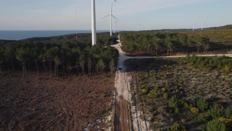 white-campervan-driving-out-of-a-forest-in-Nazare-on-a-sandy-road-lined-with-wind-turbines