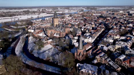Aerial-pan-around-winter-wonderland-rooftops-covered-in-snow-with-Drogenapstoren-and-historic-Hanseatic-Dutch-tower-town-Zutphen-in-The-Netherlands-with-Walburgiskerk-cathedral-and-river-IJssel-behind