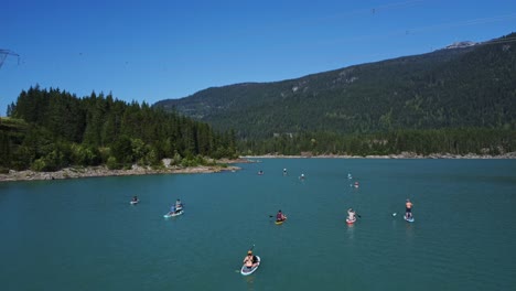 Aerial-view-approaching-a-group-of-paddle-boarders,-ascending-upwards,-flying-over-them-as-they-float-in-an-emerald-green-and-blue-Lake-surrounded-by-islands-Rocky-shoreline-forest-trees,-mountains