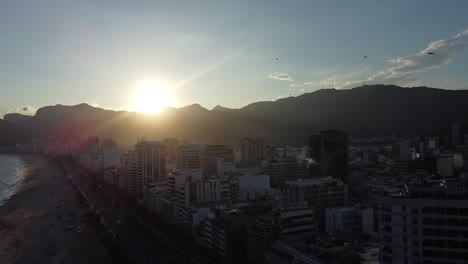 Sunset-at-Rio-de-Janeiro-with-birds-flying-in-slow-motion,-Arpoador,-with-buildings-and-mounts-backlit---aerial-shot