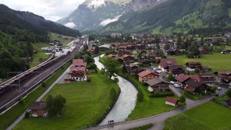 Descending-Cityscape-Aerial:-Group-of-tourist-hikers-standing-on-Bridge-by-trainStation-beside-Alpine-Glacier-kander-river-in-swiss-village-of-Kandersteg-in-Bluemlisalp-famous-for-lake-Oeschienensee