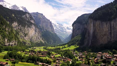 Flying-over-Lauterbrunnen-valley-town-in-Switzerland-on-a-sunny-summer-day,-aerial-view-of-famous-Swiss-alpine-village-with-Staubbach-waterfall,-snow-covered-mountain-peaks,-houses-among-green-meadows