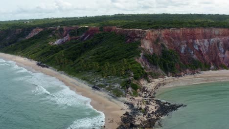 Dolly-out-aerial-drone-wide-shot-of-the-popular-tropical-Coquerinhos-beach-with-colorful-cliffs,-palm-trees,-golden-sand,-small-waves,-and-turquoise-water-in-Conde,-Paraiba,-Brazil