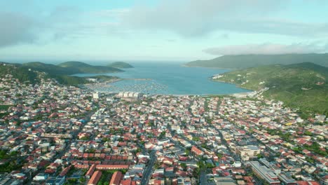 Vista-Panorámica-De-Drones-De-La-Playa-Dos-Anjos-Arraial-Do-Cabo-Brasil-Paisaje-Tropical,-Colinas-Verdes,-Barrio-Costero-Y-Horizonte-Azul-Nublado