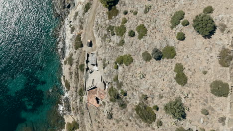 Aerial-view-looking-down-over-rustic-stone-build-house-on-Cap-de-Creus-cliffs-of-the-national-park-rocky-mountainside-and-shimmering-emerald-water