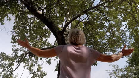 Slow-rotating-shot-of-an-elderly-woman-meditating-and-lifting-her-arms-under-a-tree