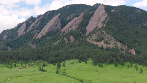 Close-up-rotating-aerial-view-of-warm-sun-hitting-Boulder-Colorado-Flatiron-mountains-above-Chautauqua-Park-with-full-green-pine-trees-and-blue-skies-with-clouds-on-a-beautiful-summer-day-for-hiking
