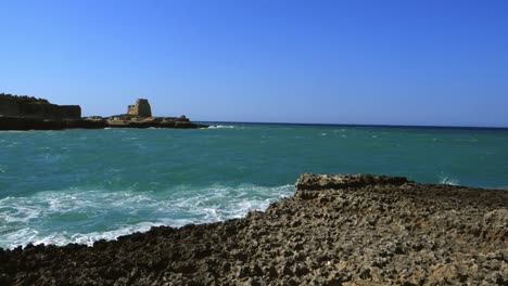 Waves-hitting-rocks-on-Ionian-sea,-an-old-tower-in-the-background
