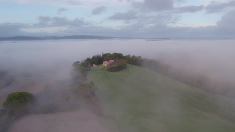 Hill-with-farm-house-sticking-out-of-mist-in-Italy,-aerial