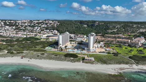 A-cinematic-aerial-view-of-a-the-buildings-with-Son-Bou-Beach-in-Menorca,-Spain
