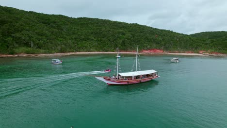 Touristic-boat-near-the-Tartaruga-beach-shoreline-In-Brazil,-ships-gracefully-navigate-the-waters-while-the-lush-forest-stands-tall-along-the-shoreline