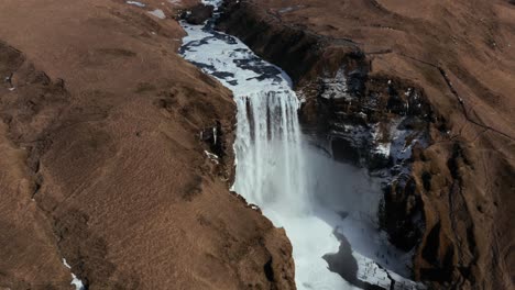 Skogafoss-waterfall-semi-frozen-surface-at-beginning-of-spring-time,-aerial