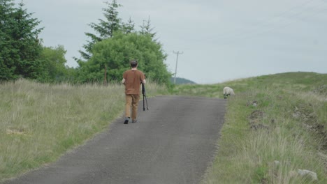 Man-using-camera-on-tripod-walking-towards-sheep-on-side-of-single-track-road