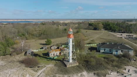Aerial-establishing-view-of-white-colored-Pape-lighthouse,-Baltic-sea-coastline,-Latvia,-white-sand-beach,-large-waves-crashing,-sunny-day-with-clouds,-orbiting-drone-shot